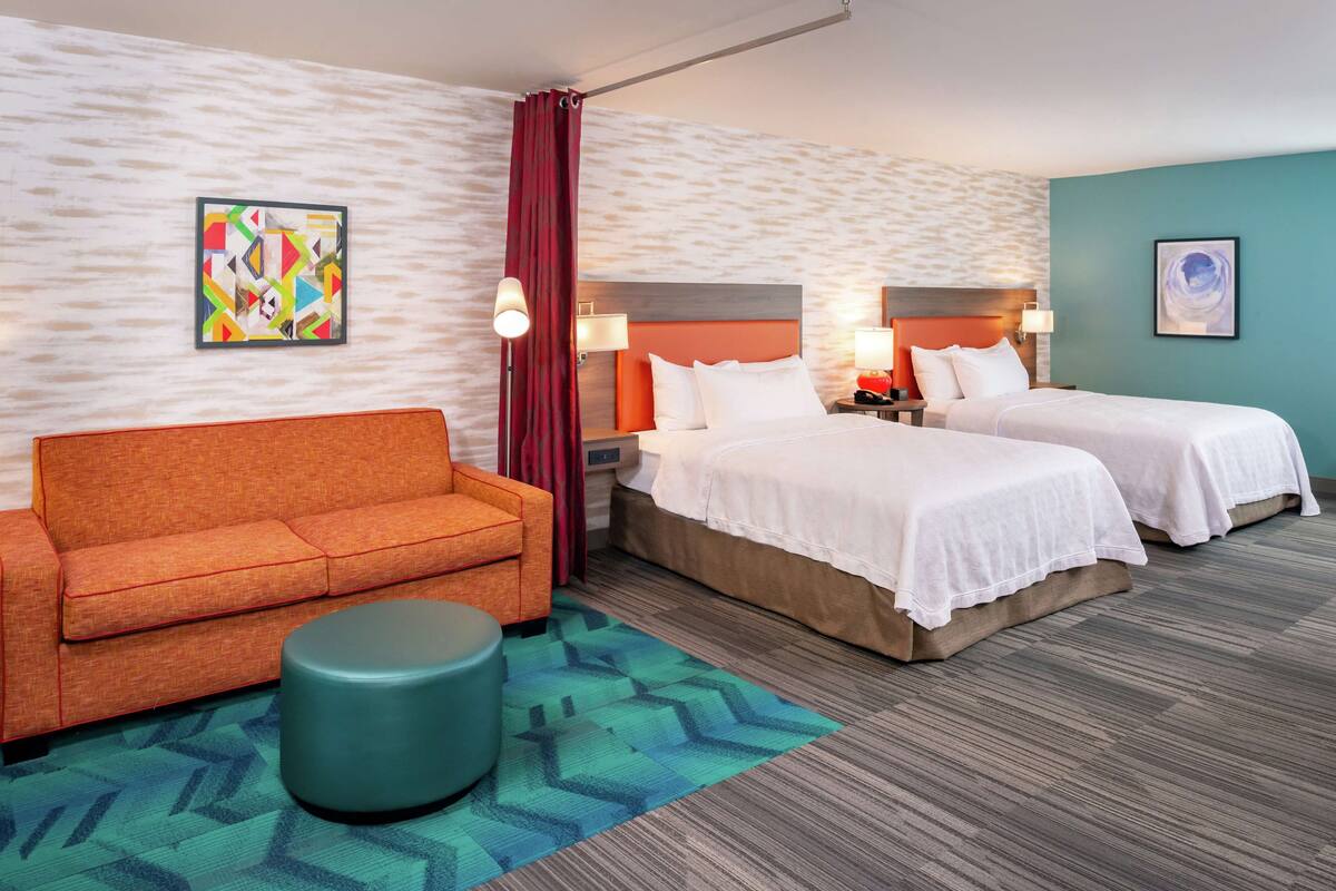 A hotel room with two beds, a sofa, a round ottoman, and artwork on the walls.