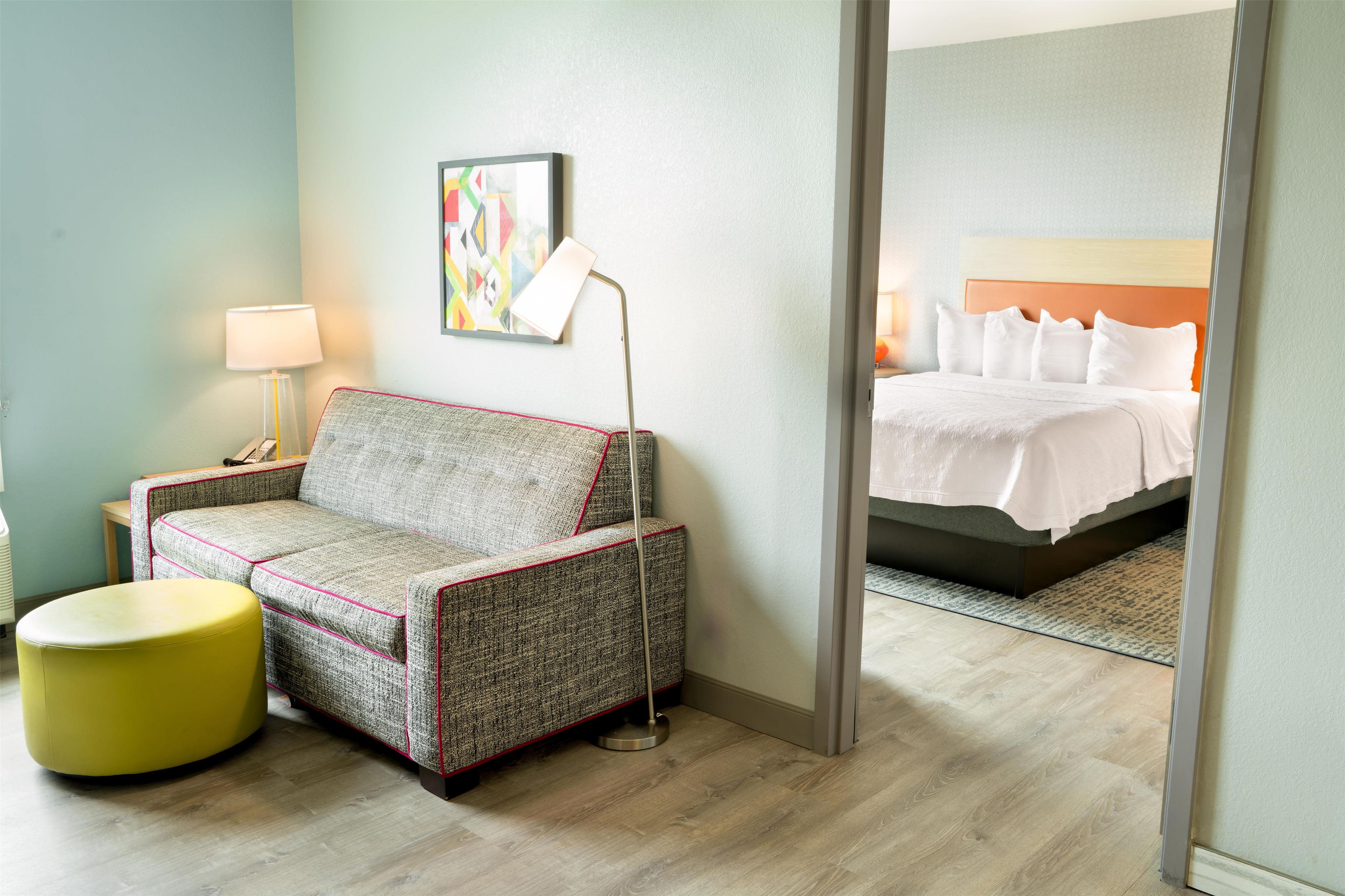 Quarto de hotel com sofá, um banquinho amarelo pequeno, luminária de chão e cama com roupa de cama branca.