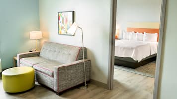 Quarto de hotel com sofá, um banquinho amarelo pequeno, luminária de chão e cama com roupa de cama branca.