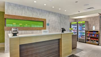Ein Hotel-Rezeptionsbereich mit einem hölzernen Empfangstresen, einer digitalen Check-in-Kiosk, einem Fenster mit grünem Rahmen sowie Verkaufsautomaten mit den Aufschriften 'wirklich kalt' und 'kalt'.