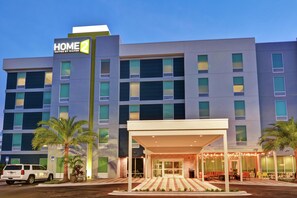 A multi-story hotel with a prominent 'HOME 2' sign, a covered entrance with outdoor seating, and a parking area with a white SUV. - Home2 Suites by Hilton Jacksonville-South/St. Johns Town Ctr (Jacksonville)