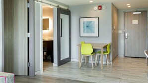 A hotel room with a wooden floor, a dining area with a table and chairs, a bathroom with a towel rack, and a door leading to another room. - Home2 Suites by Hilton Jacksonville-South/St. Johns Town Ctr (Jacksonville)