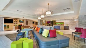 A modern hotel lobby with a large seating area, a reception desk, and a kitchen visible in the background. - Home2 Suites by Hilton Jacksonville-South/St. Johns Town Ctr (Jacksonville)