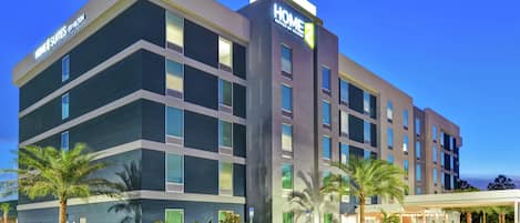 Un hôtel à plusieurs étages, doté d’un design moderne, qui se distingue par une grande enseigne « HOME », une entrée abritée et un parking où sont stationnés plusieurs véhicules.