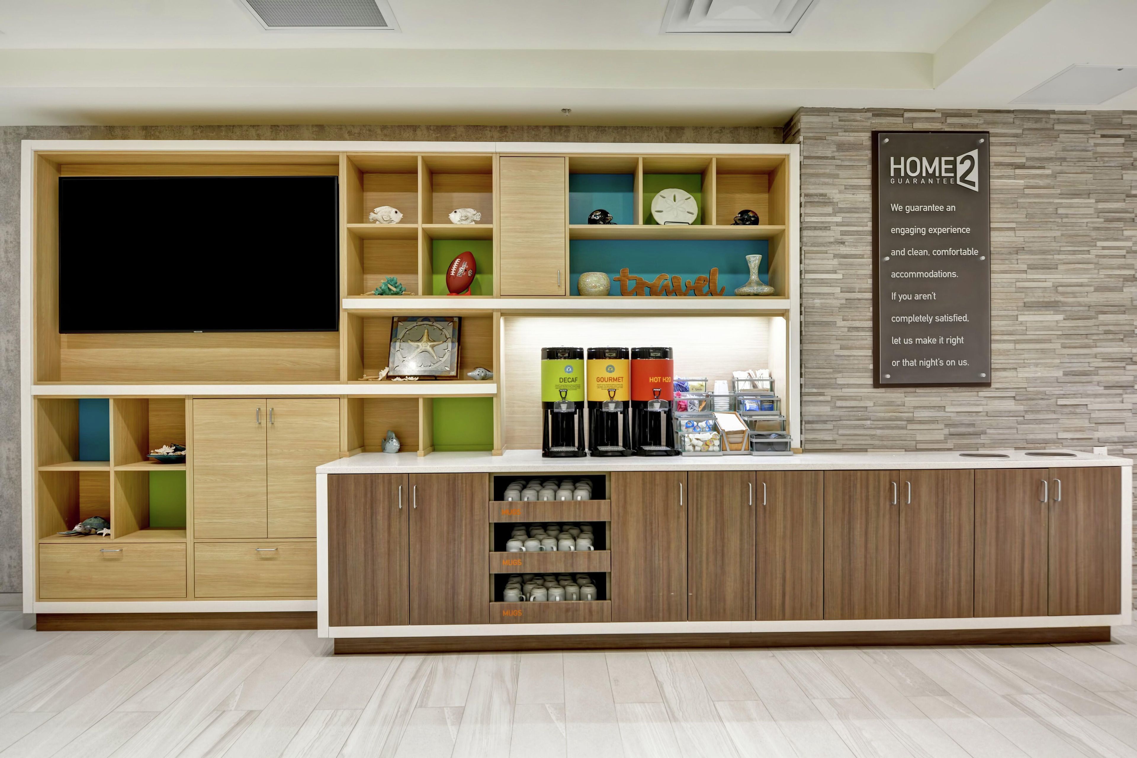A modern hotel lobby with a large flat-screen TV, a coffee station, and a sign stating 'HOME2 GUARANTEE'.