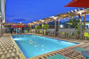 A pool area with a tiled deck, a pool with a depth marker, and outdoor seating with umbrellas. - Home2 Suites by Hilton Jacksonville-South/St. Johns Town Ctr (Jacksonville)
