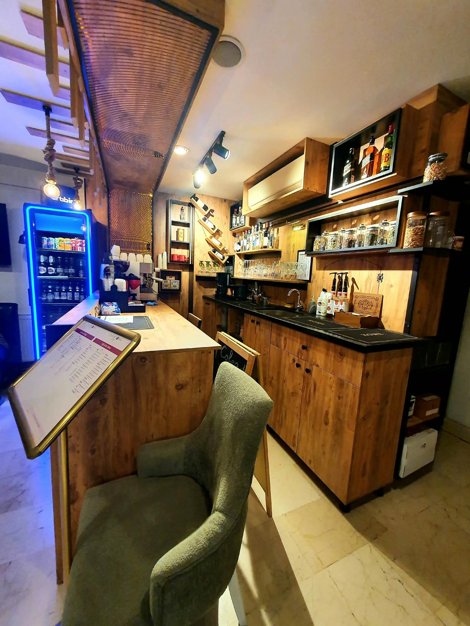 A bar area with a wooden counter, a seating area with a chair, and a refrigerator stocked with beverages.