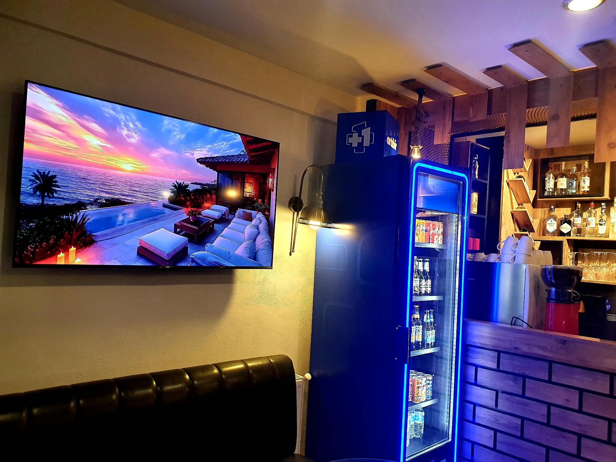 A bar area with a TV displaying a sunset view, a blue illuminated refrigerator, and a brick wall.