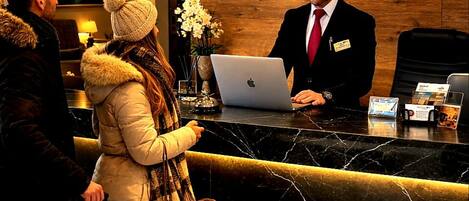 A hotel reception desk with a receptionist, a laptop, and guests with luggage.