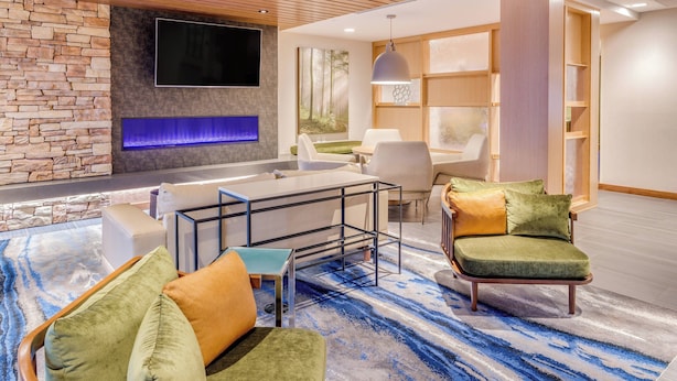 A modern living room with a stone wall, a flat-screen TV, a blue-lit fireplace, a dining area with a table and chairs, and a large window with a forest view. - Fairfield Inn & Suites Jasper (Jasper)