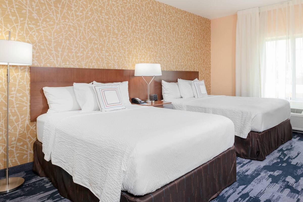 A hotel room with two beds, a wooden headboard, a patterned wall, and a large window with curtains.