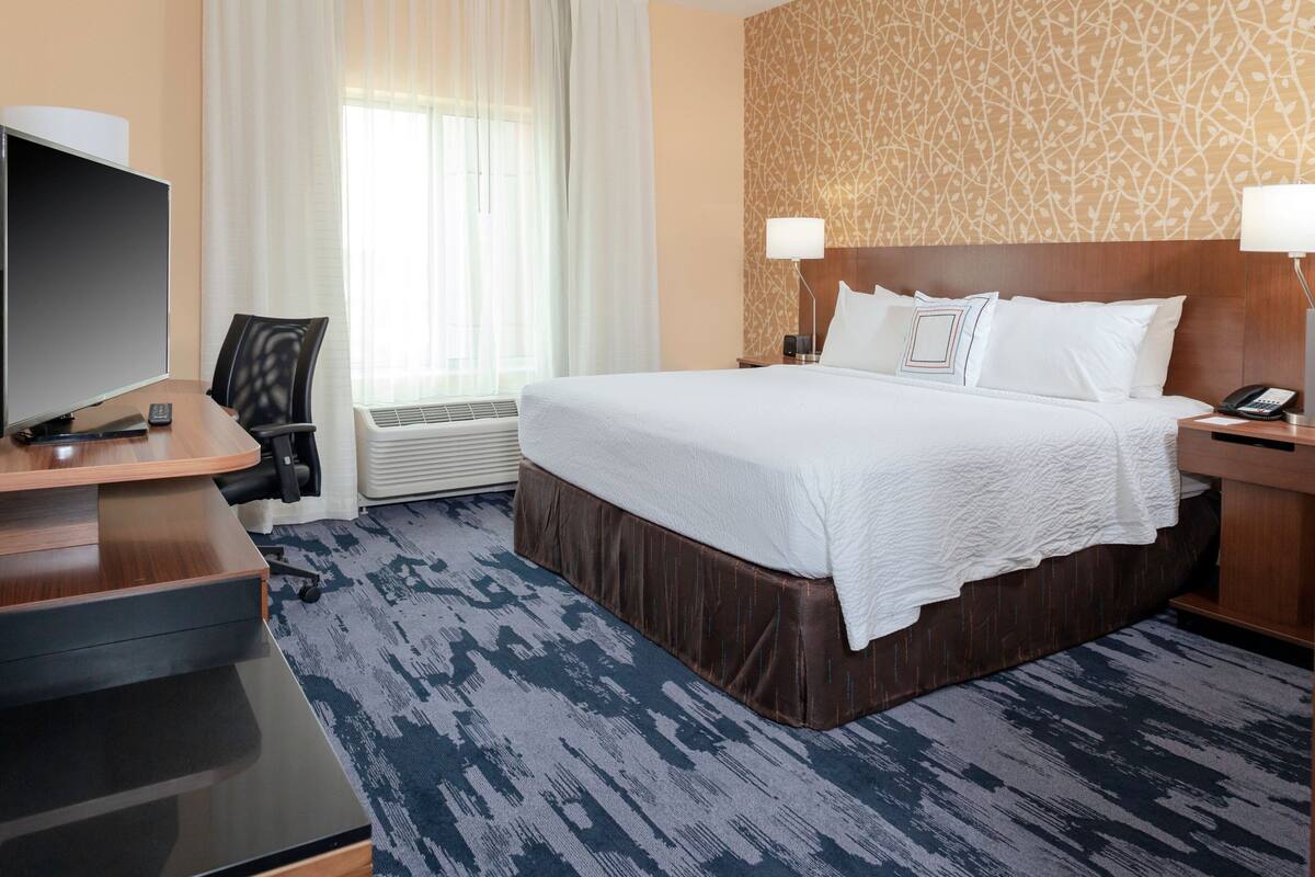 A hotel room with a large bed, a desk with a flat-screen TV, a chair, a nightstand with a lamp, and a patterned wall.