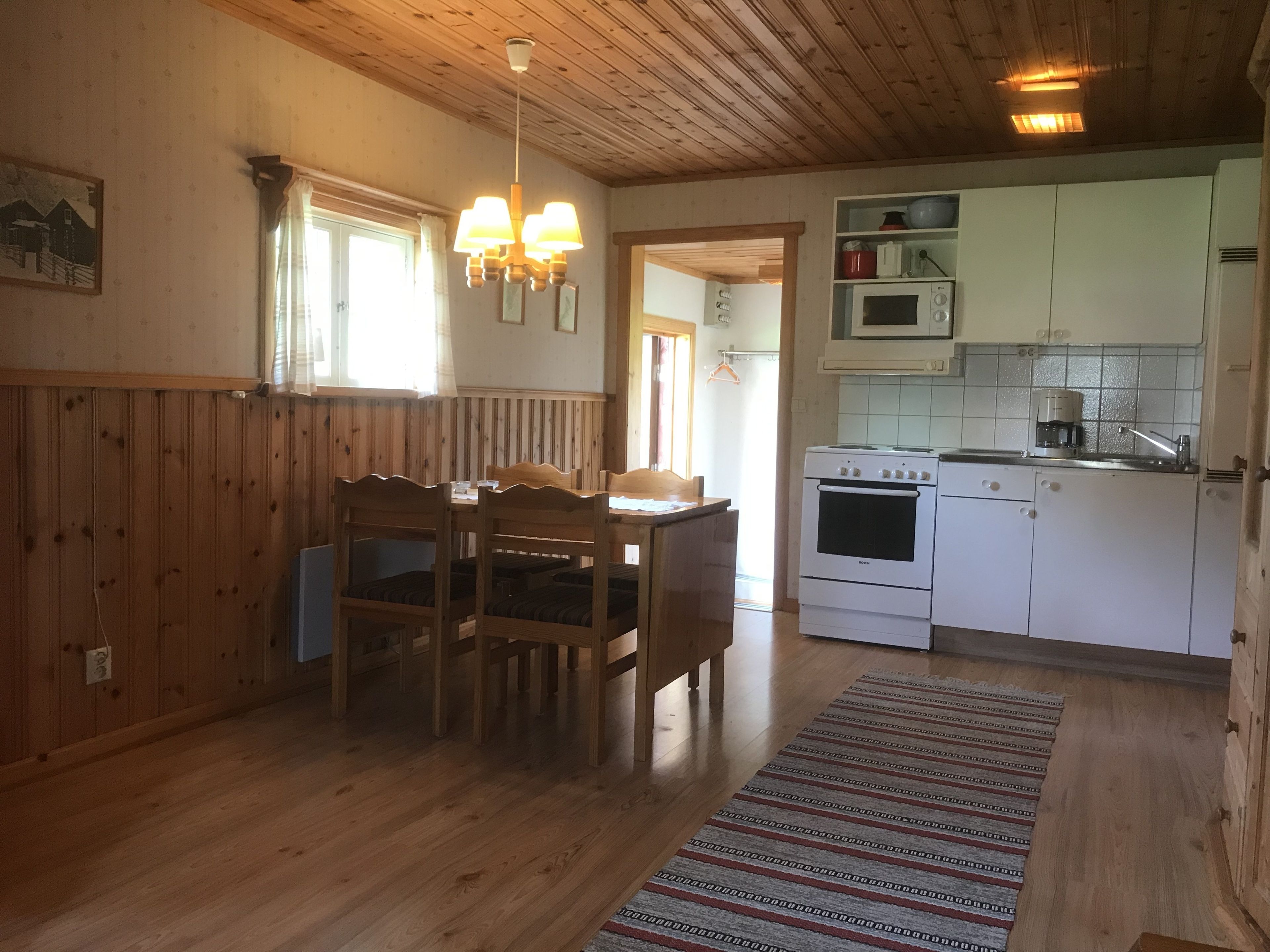 A wooden-paneled room with a dining table and chairs, a kitchen with white cabinets and appliances, and a patterned rug on the floor.