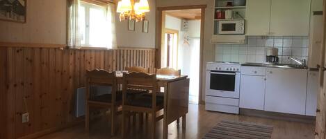 A wooden-paneled room with a dining table and chairs, a kitchen with white cabinets and appliances, and a patterned rug on the floor.
