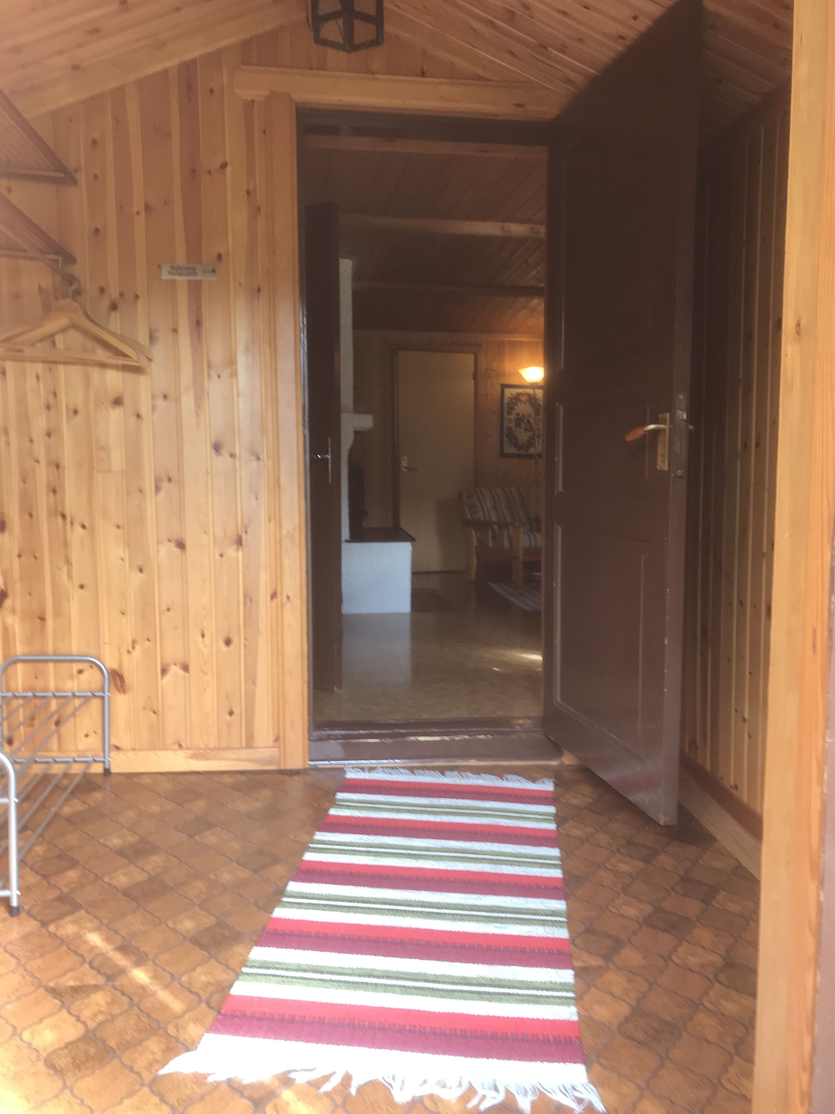 A wooden interior with a hallway leading to a room. A striped rug is on the floor, and a metal chair is visible.
