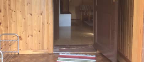 A wooden interior with a hallway leading to a room. A striped rug is on the floor, and a metal chair is visible.