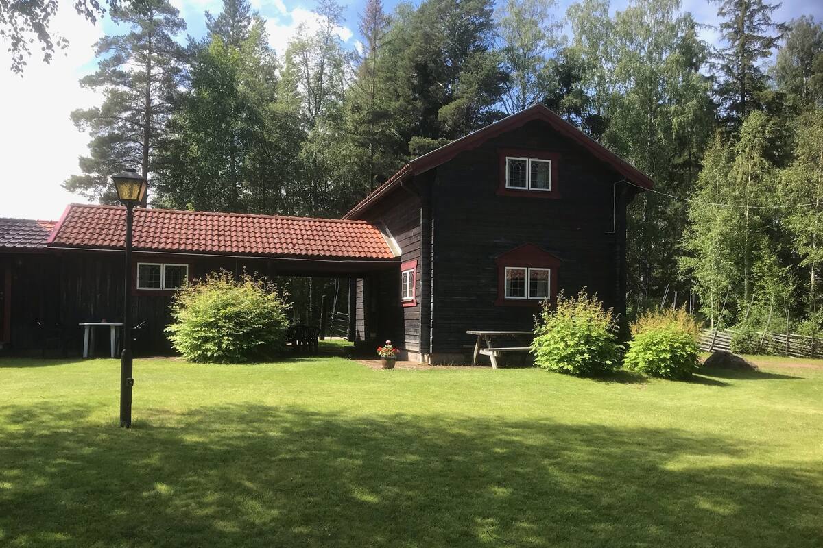 A two-story wooden house with a red-tiled roof, a covered porch, and a small garden area with a table and chairs.