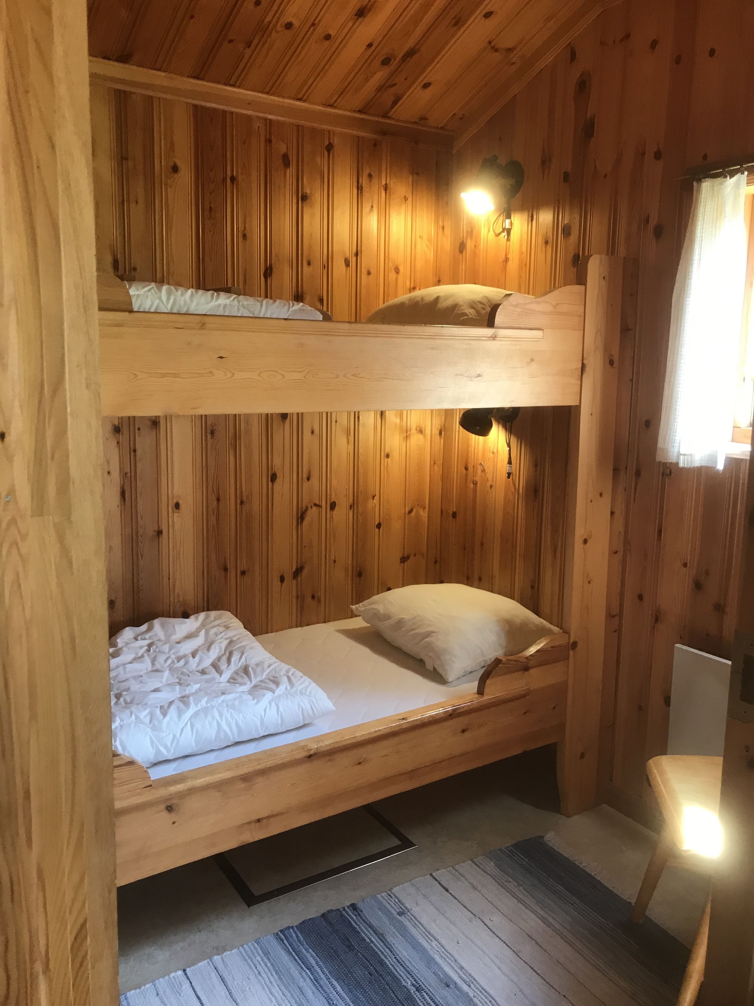 Ein Stockbett mit zwei Etagen, Holzvertäfelung, ein kleines Fenster mit Vorhängen und ein Stuhl.
