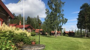 Hyvin hoidettu nurmikko, jonka keskellä on pyöreä kivipöytä ja ruukussa oleva kasvi. Kaikkea tätä ympäröi punaisella katolla varustettu, mökin tyylinen puurakennus.