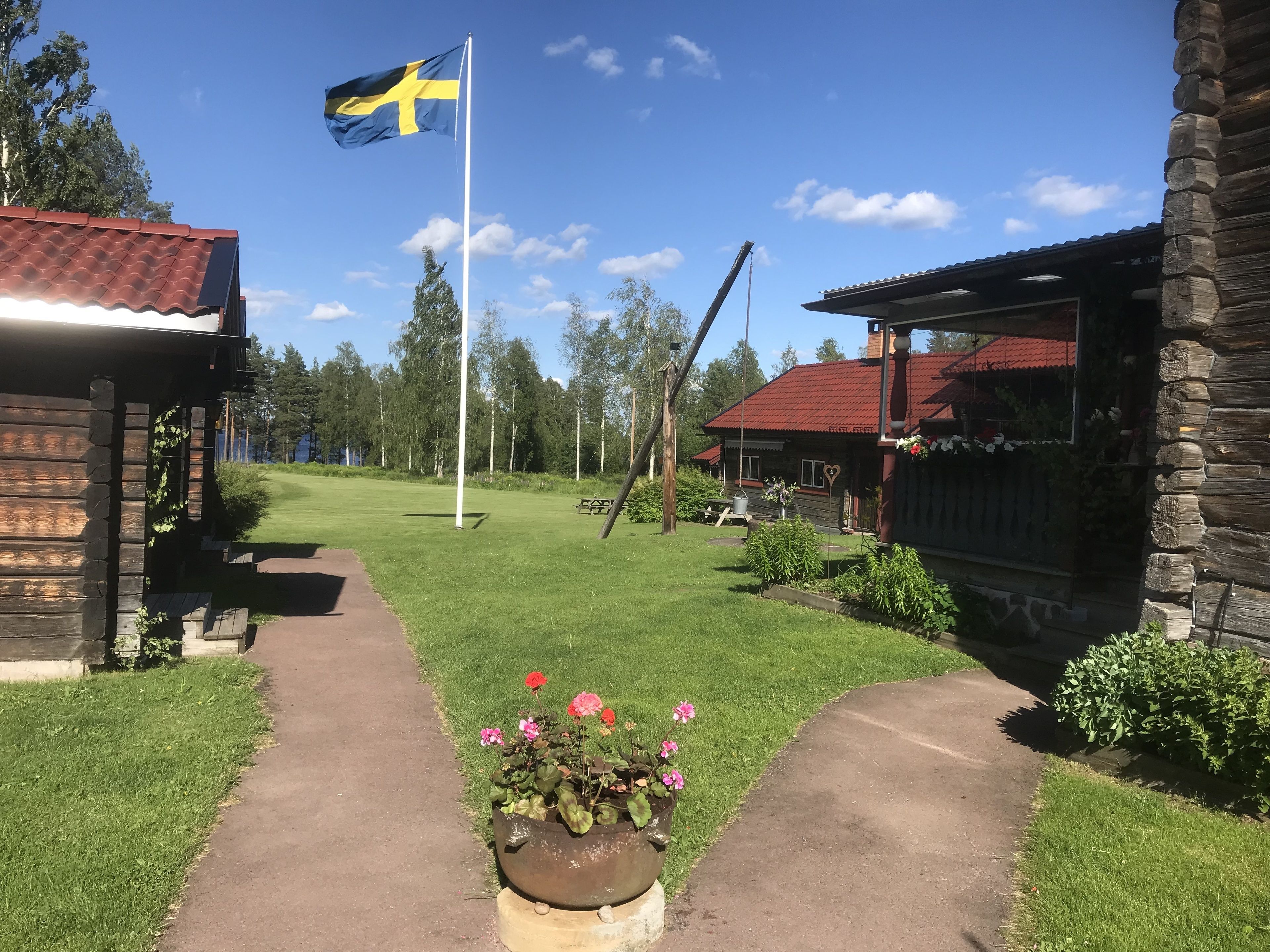 A wooden cabin with a red roof, a flagpole with a flag, and a pathway leading to a grassy area with flower beds.