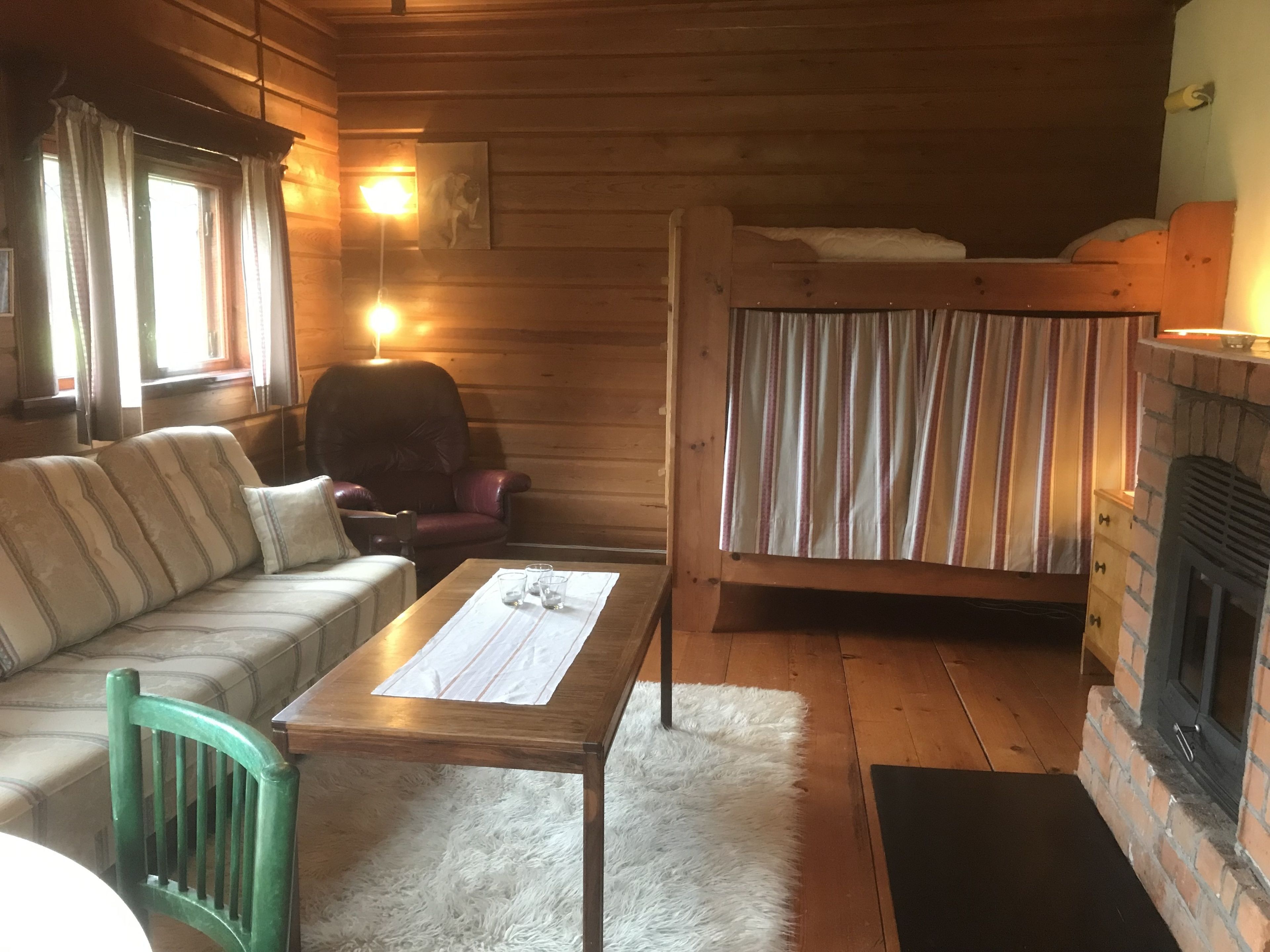 A cozy cabin interior with a wooden floor, a sofa, a green chair, a table with glasses, a bunk bed, and a fireplace.