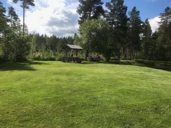 A well-maintained grassy area with a wooden gazebo and picnic tables, surrounded by dense trees and a clear sky. - Tollagården (Sollerön)
