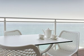 A white table with a teapot, cups, and saucers on a balcony with a view of the sea.