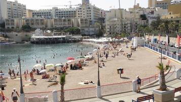 A beach with people sunbathing and swimming, a promenade with benches, and buildings in the background.