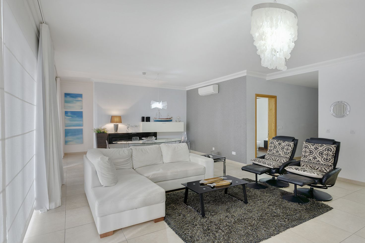 A modern living room with a white sofa, a coffee table, and two black armchairs. There is a kitchen area in the background with a white countertop and a hanging light fixture.