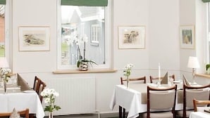 Ein Essbereich mit Tischen für vier Personen, einer Kristallleuchter, gerahmten Kunstwerken an den Wänden und Blick durch ein Fenster mit grünen Vorhängen.