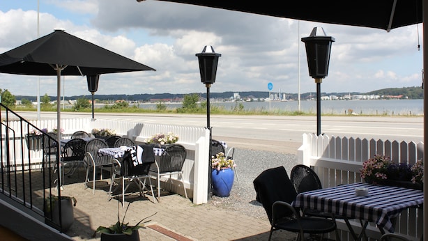 Eine Terrasse im Freien mit Tischen und Stühlen, Blick auf ein Gewässer und ein weißes Zäunchen.