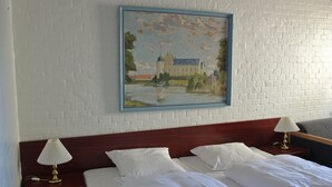 Ein Hotelzimmer mit einem Bett, zwei Nachttischen mit Lampen, einem Gemälde eines Schlosses an der Wand und einer weißen Ziegelwand.