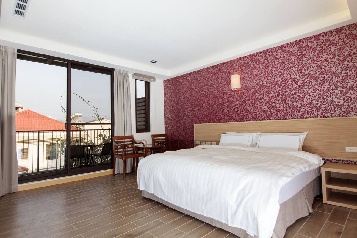 A hotel room with a large bed, a wooden headboard, a nightstand, and a balcony with chairs.