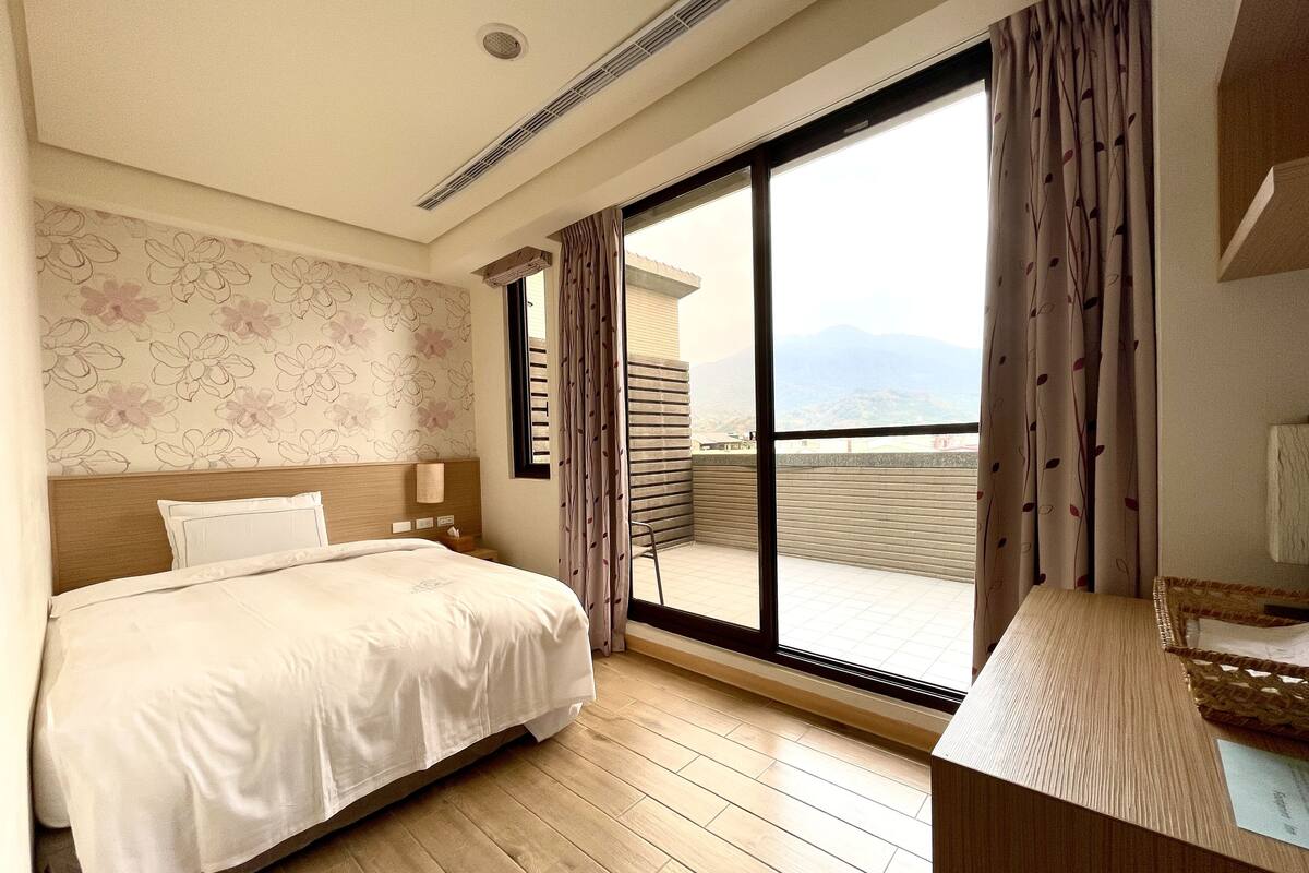 A hotel room with a bed, a bedside table, a mirror, a window with curtains, and a view of the mountains.