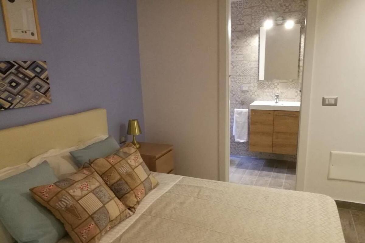 A bedroom with a bed, pillows, a bedside table, and a nightstand. There is a bathroom visible through an open door.