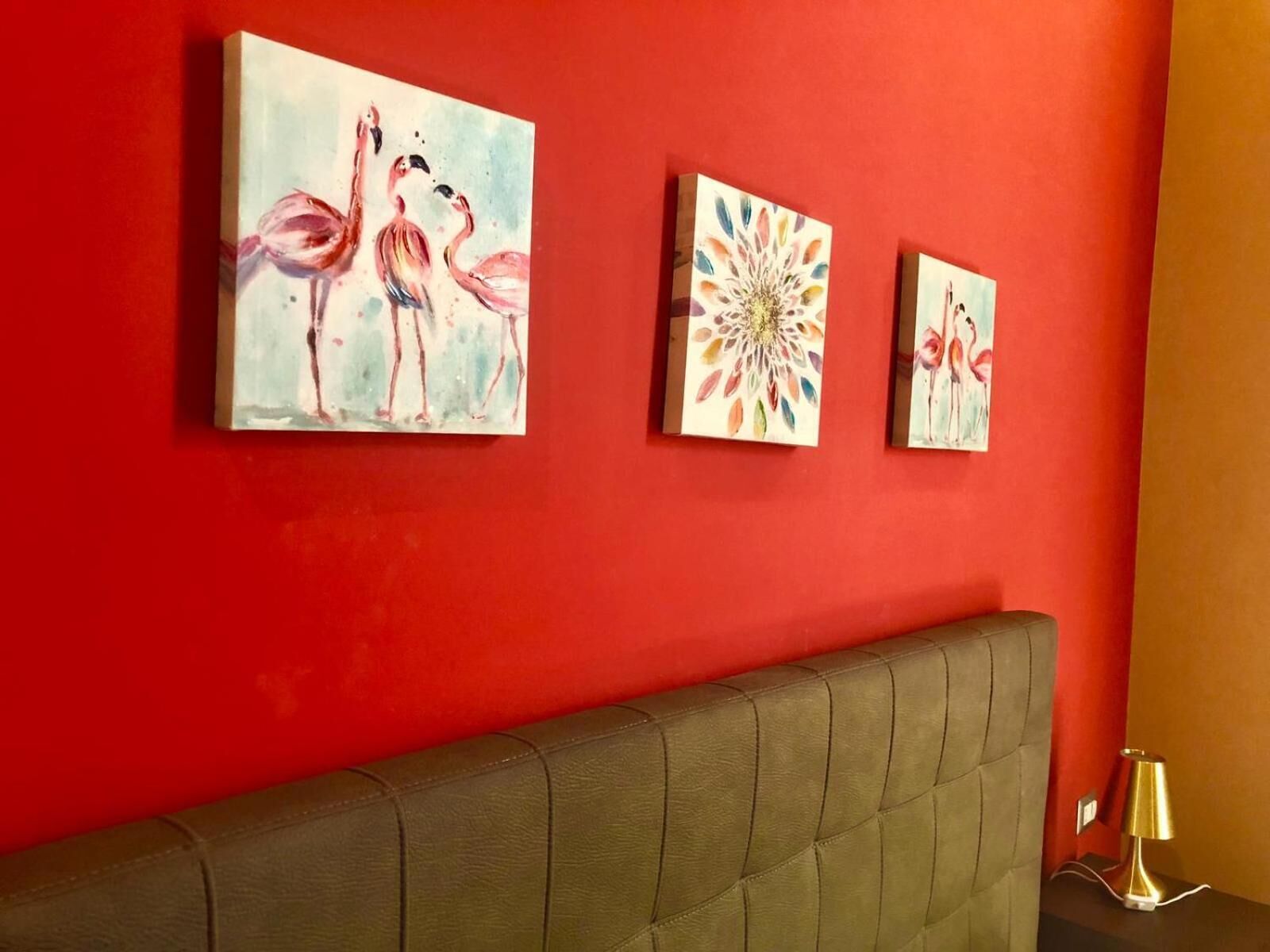 A room with a red wall, a brown headboard, and three paintings: two of flamingos and one of a colorful flower.