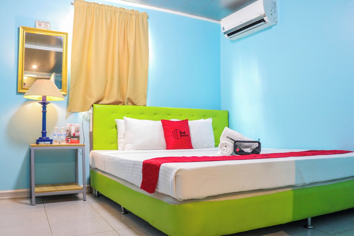 A room with a green bed, white bedding, a red pillow, a small table with a lamp, and an air conditioning unit.