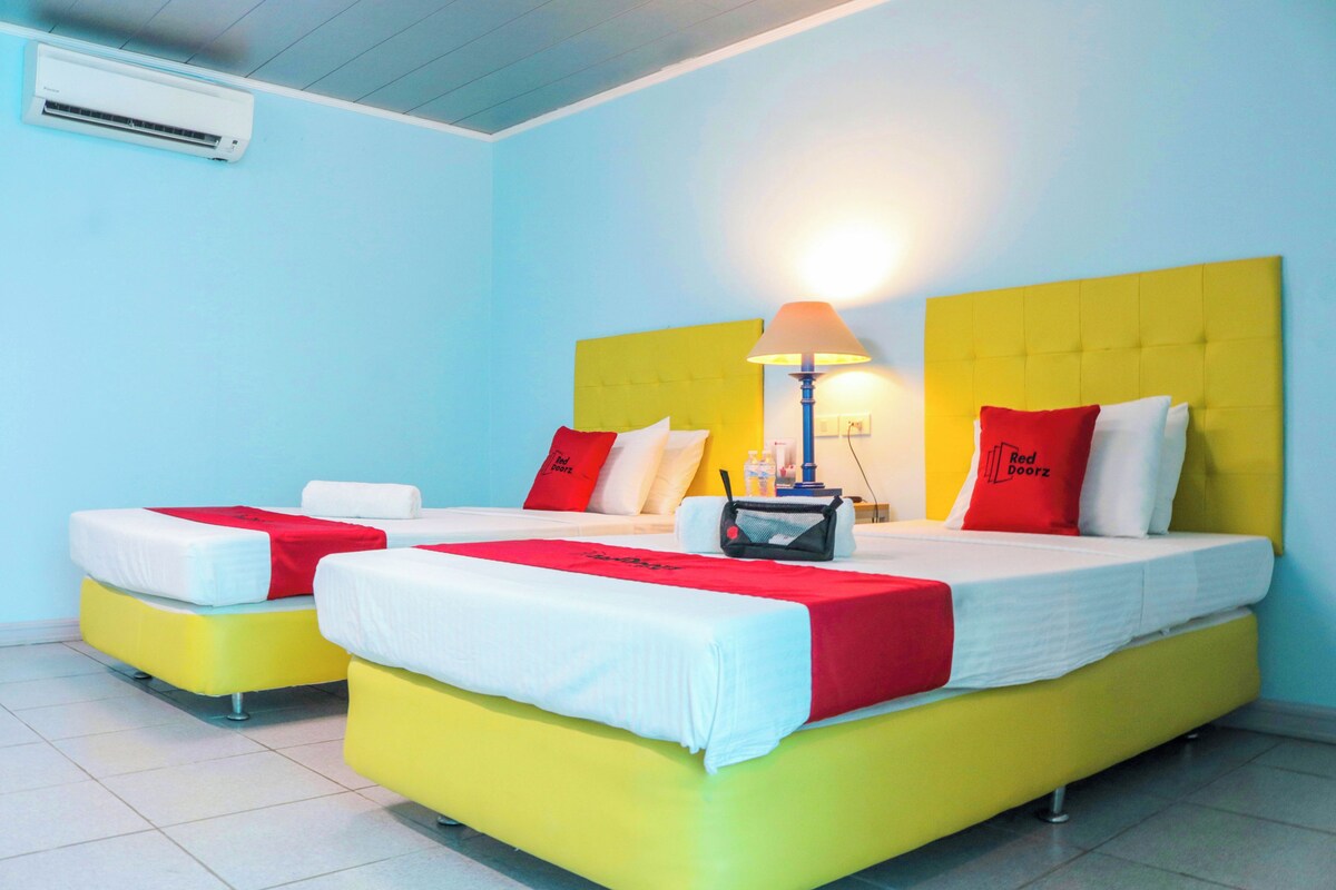 A hotel room with two beds, each with a yellow headboard and white bedding. There is a lamp on the nightstand, and an air conditioning unit on the wall.