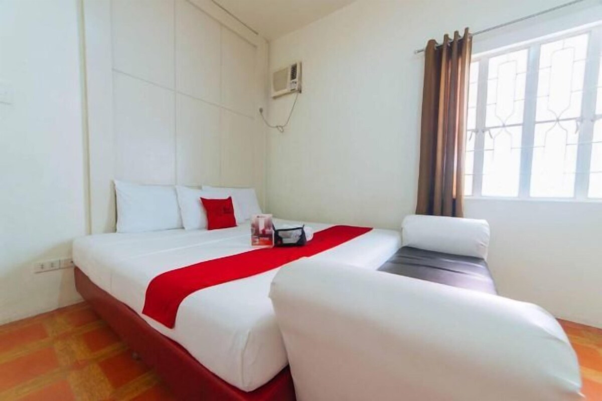 A hotel room with a bed, a chair, a window with curtains, and an air conditioning unit.