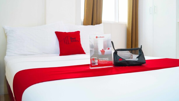Habitación de hotel con una cama, una almohada roja, un cartel promocional de RedDoorz y una bolsa negra sobre la cama.