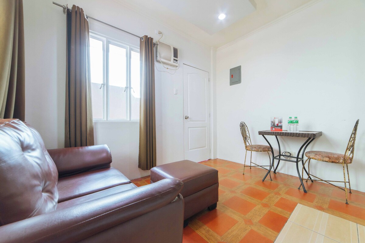 Une pièce avec un canapé en cuir marron, une petite table avec deux chaises et une fenêtre avec des rideaux.