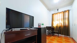 Habitación con televisión de pantalla plana sobre un mueble de madera, un refrigerador pequeño, una silla y una ventana con cortinas.