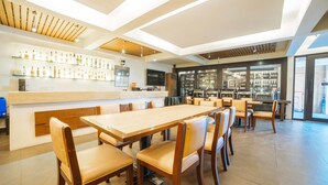 Un restaurante moderno con una larga mesa de madera y sillas acolchadas, una zona de bar con estantes llenos de botellas y una cocina al fondo.