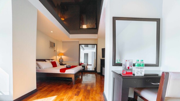 Habitación de hotel con cama, mesita de noche, lámpara y escritorio con silla. Se observa un espejo en la pared y una puerta que da a otra habitación.