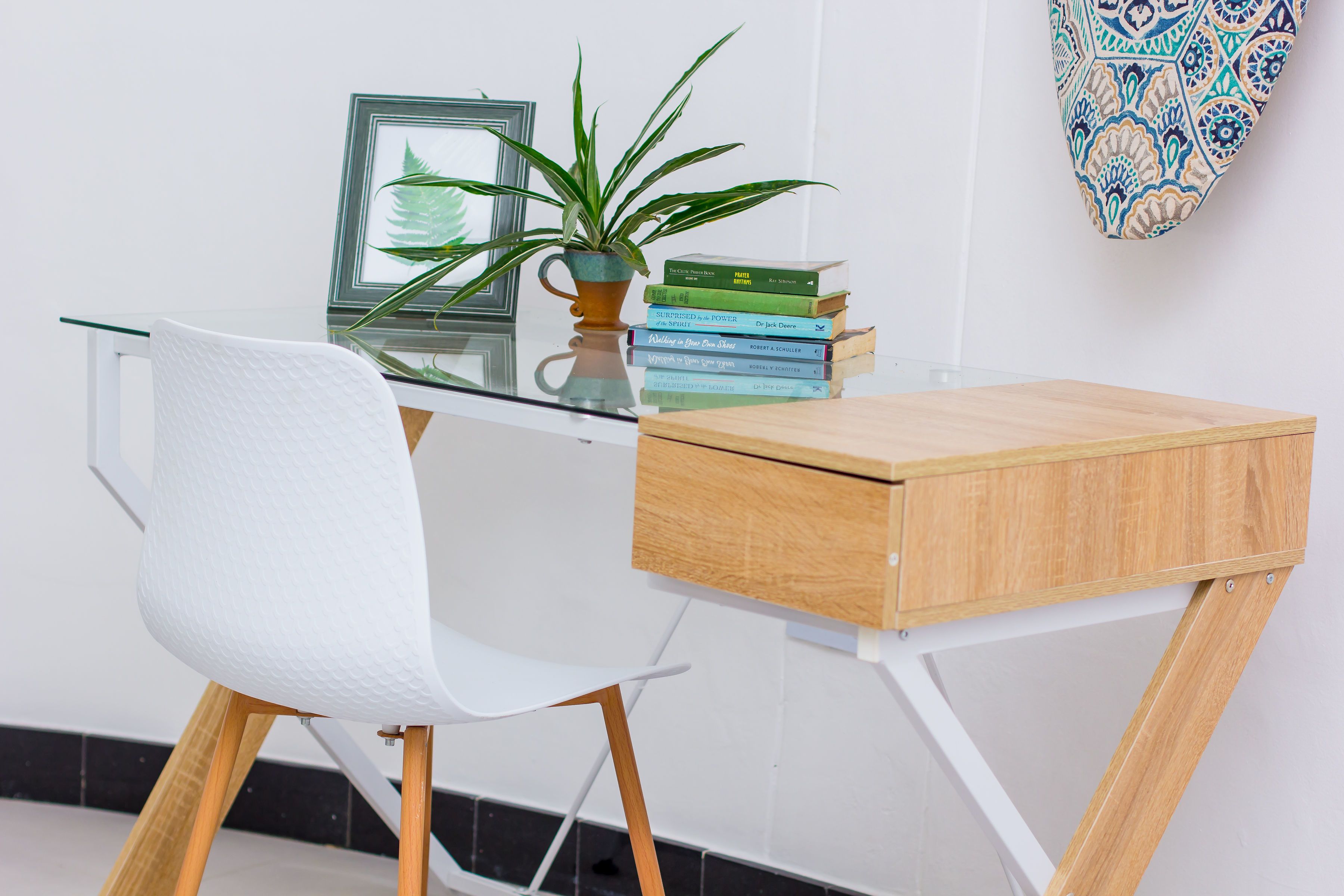 A modern workspace with a glass desk, white chair, wooden drawer, and a potted plant.