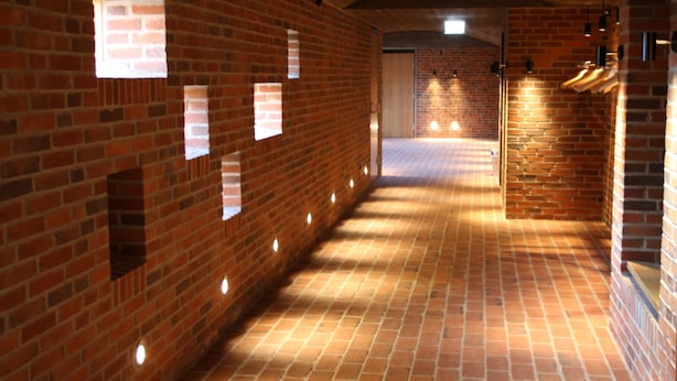 Un pasillo con paredes de ladrillo, iluminación empotrada, una puerta de madera al final y una pared de ladrillo a la derecha con luces colgantes.