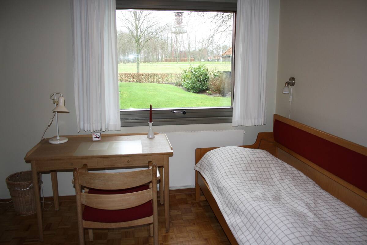 Ein Zimmer mit einem Holztisch, einem Stuhl, einem Bett mit rotem Kopfteil und einem Fenster mit Blick auf eine Wiese und Bäume.