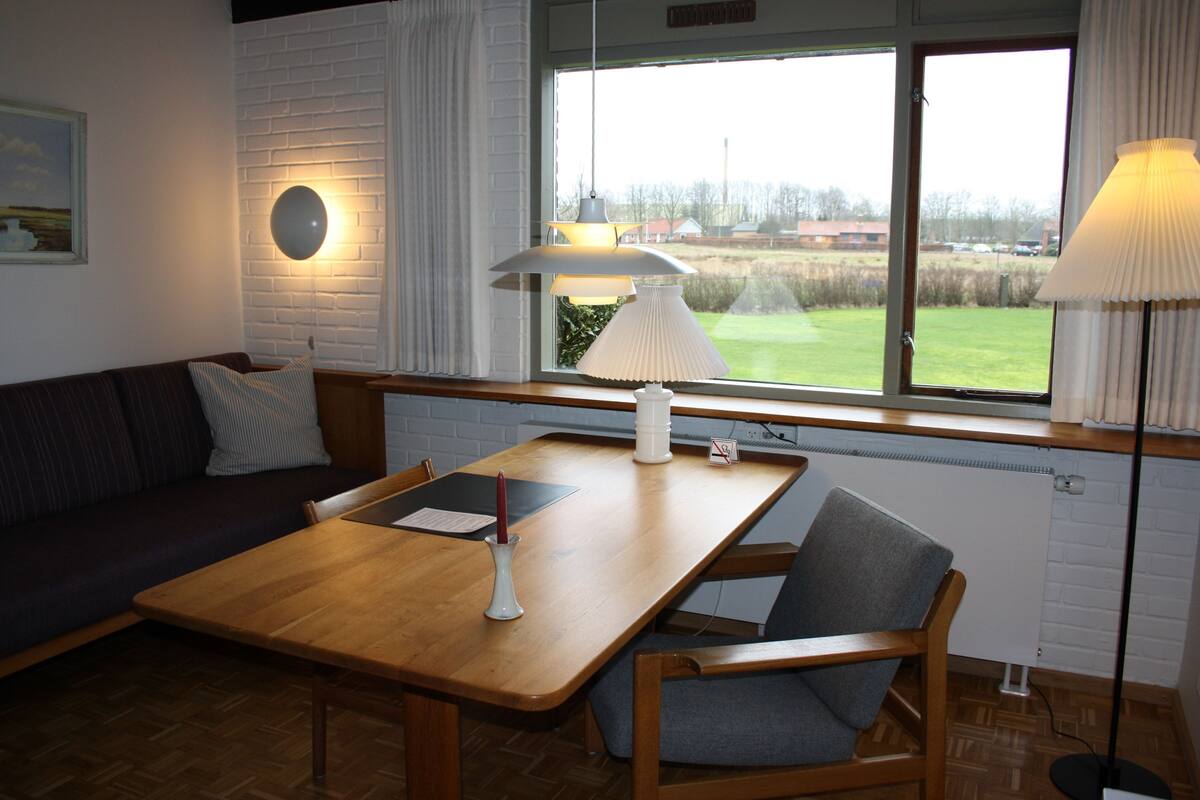 Ein gemütliches Zimmer mit einem Holztisch, einer gepolsterten Bank und Blick auf ein grünes Feld durch ein großes Fenster.
