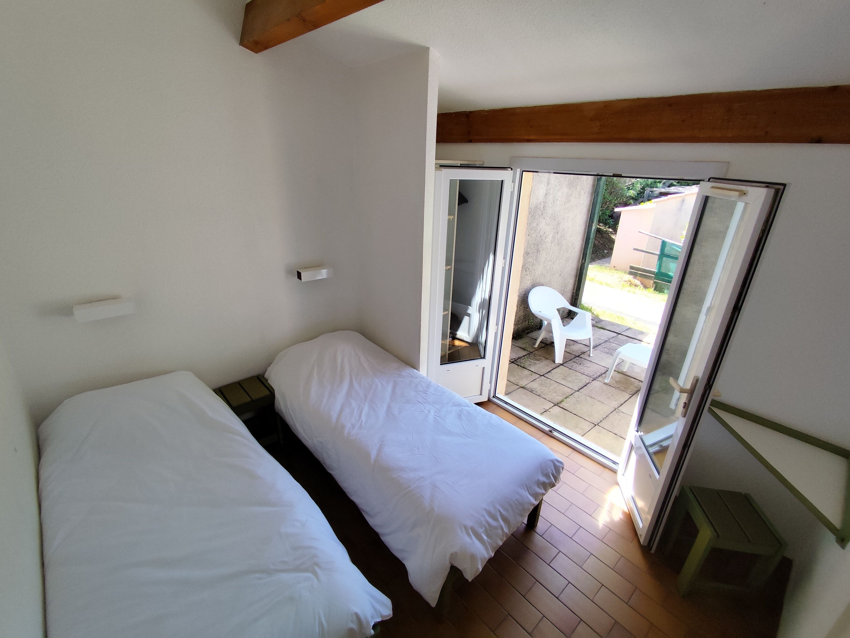 Ein kleines Zimmer mit einem Einzelbett und einer Schiebetür zu einem Außenbereich mit Terrasse und einem weißen Stuhl.