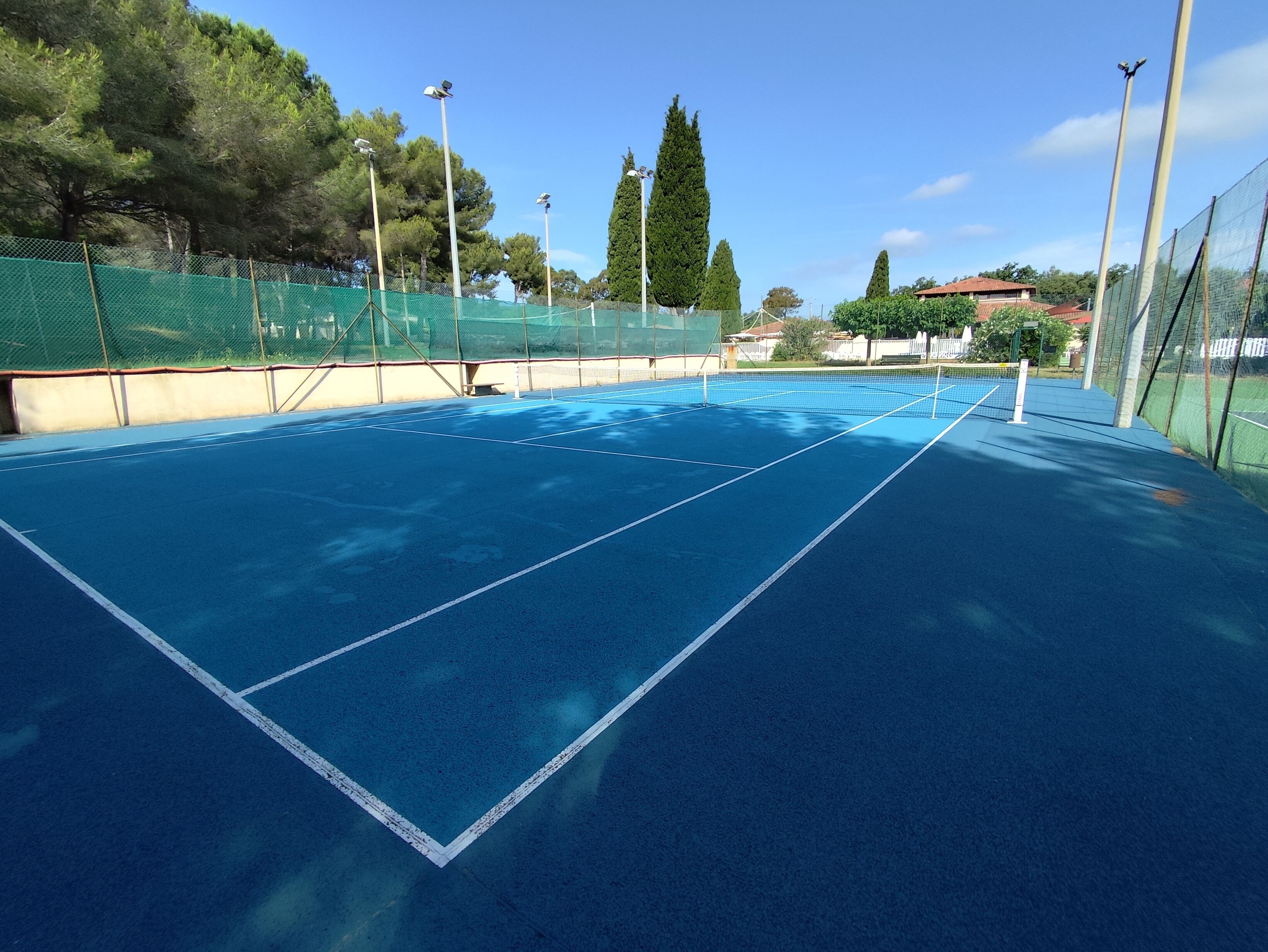 Un campo da tennis con superficie blu, circondato da recinzioni verdi e alberi alti.